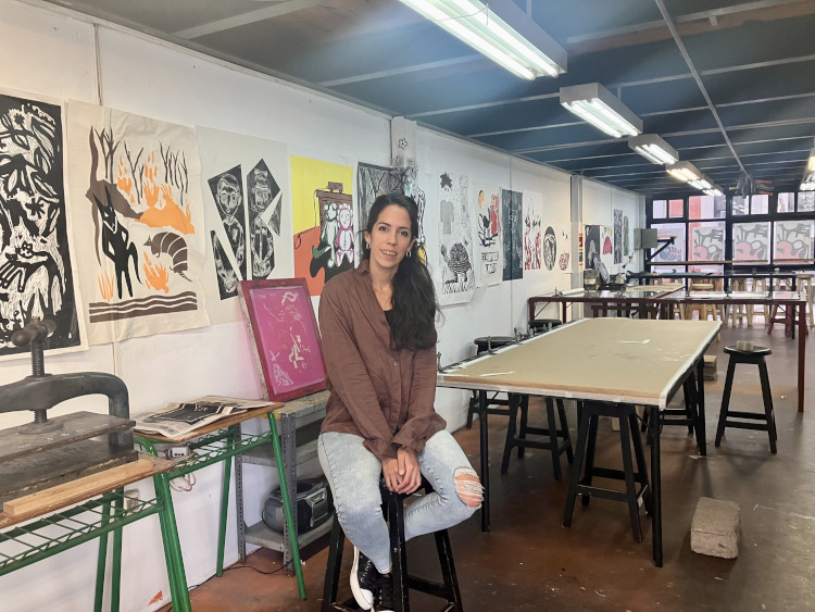 Foto de Martina Mansilla sentada en el salón del taller de Artes Gráficas de Facultad de Artes. Ventanal detrás y afiches de serigrafía pegados en la pared. 
